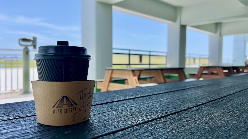 道の駅かでなの展望デッキの木製テーブルに置かれたALTO COFFEEのカップと、背景に広がる嘉手納基地の滑走路