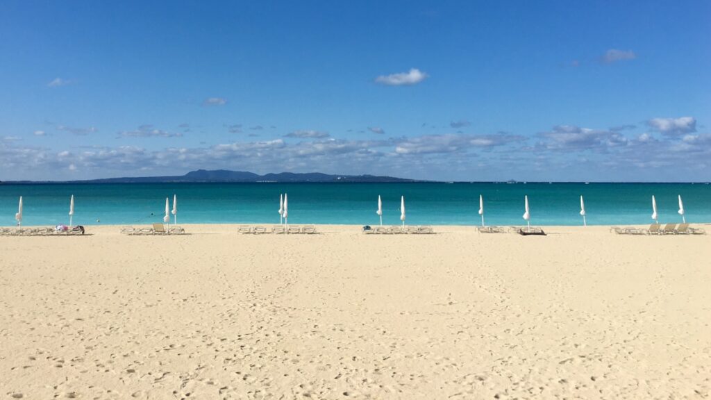 青空とエメラルドグリーンの海を背景に白い砂の上に、白い閉じられたパラソルとビーチベッドが並ぶ沖縄のオクマビーチの風景