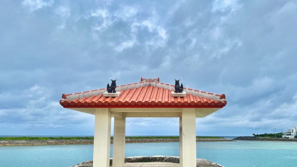 曇り空と青い海を背景に、赤瓦屋根にシーサーが鎮座する東屋がある沖縄の梅雨の風景