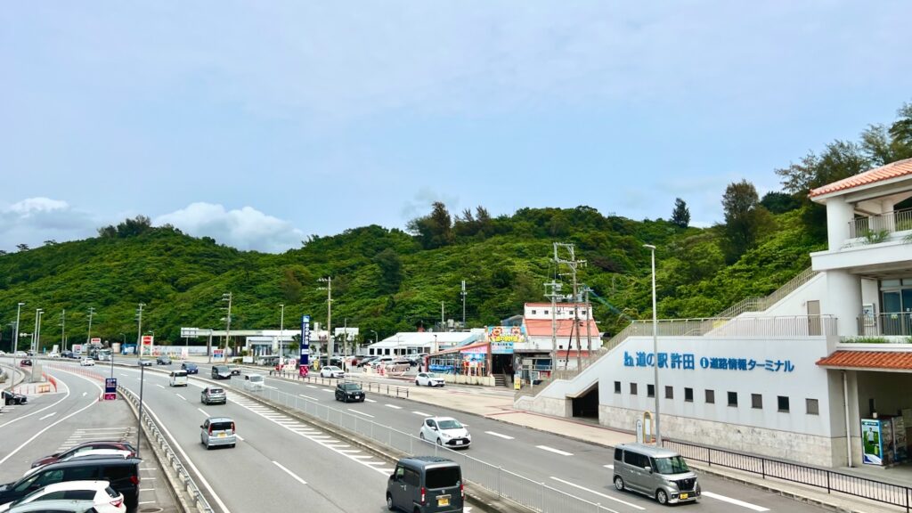 沖縄の青空と緑豊かな山々を背景にした「道の駅許田」の外観と、その前を通る道路