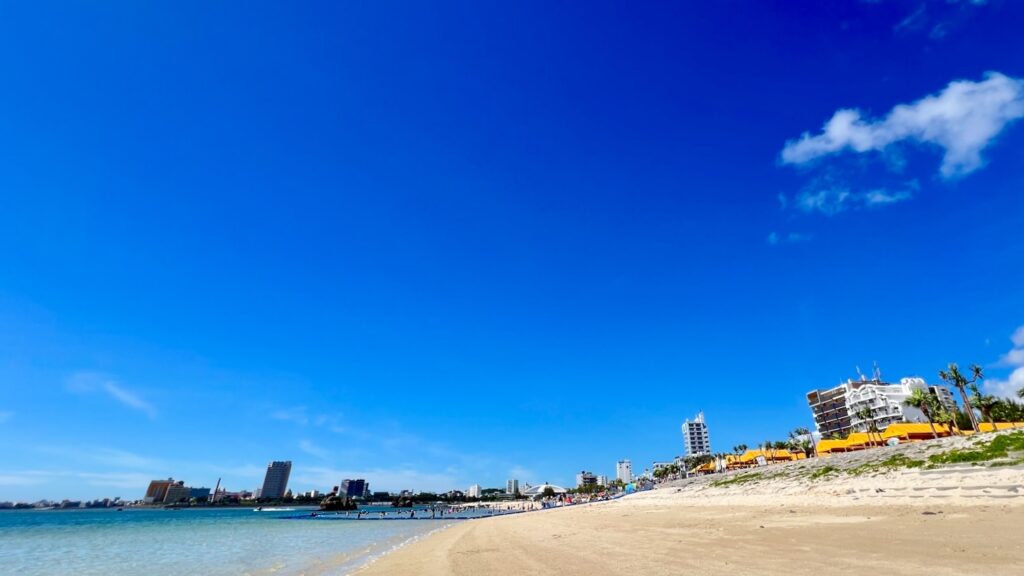 青空と北谷の建物を背景に、海と白い砂浜が広がるアラハビーチの風景