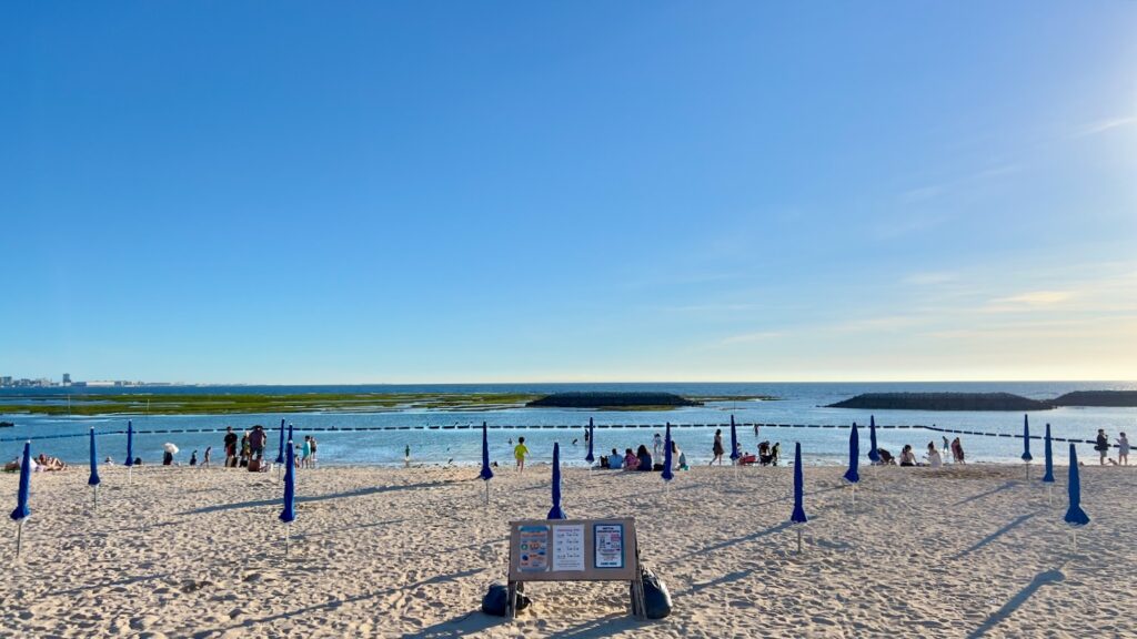 綺麗な青空と海を背景に、閉じられた青いパラソルが並ぶサンセットビーチと、ビーチを楽しむ人々