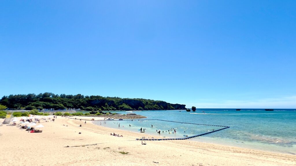 白い砂浜に白いビーチパラソルが並び、青空と木々の緑、青い海が広がる沖縄のビーチの風景