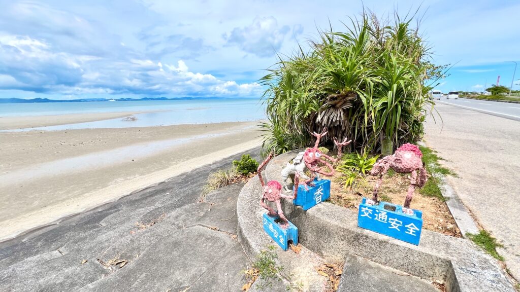 干潮時の海中道路の風景と道中に設置された、足元のブロックに「香」「交通安全」の文字がついている三体の手作りのキジムナー像