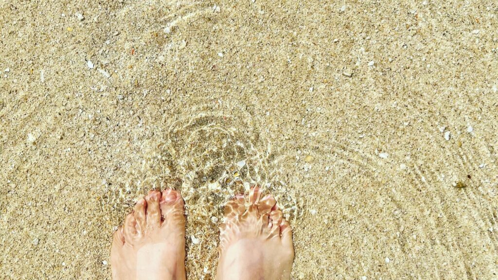 沖縄のビーチの波打ち際で、透明な海水が足元を洗う様子。細かい砂とサンゴのかけらが混ざる砂浜。