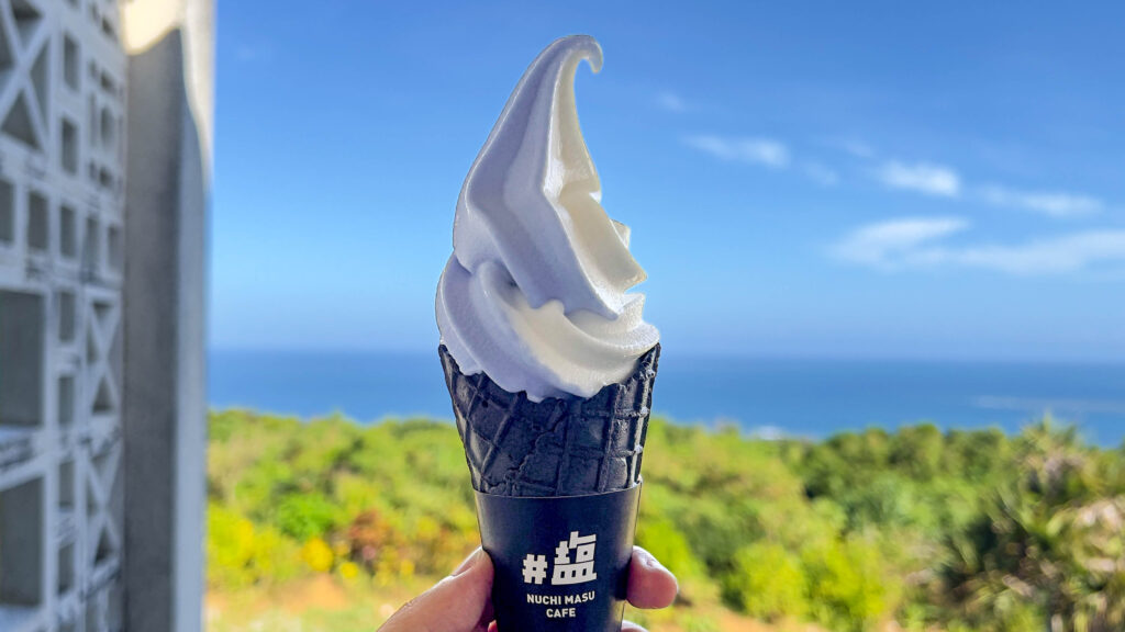 青い空、青い海、緑の植物を背景に、ぬちまーすカフェの水色と白色のソフトクリームと黒いコーンの色合いが印象的な「うみそらソフト」を手に持った画像
