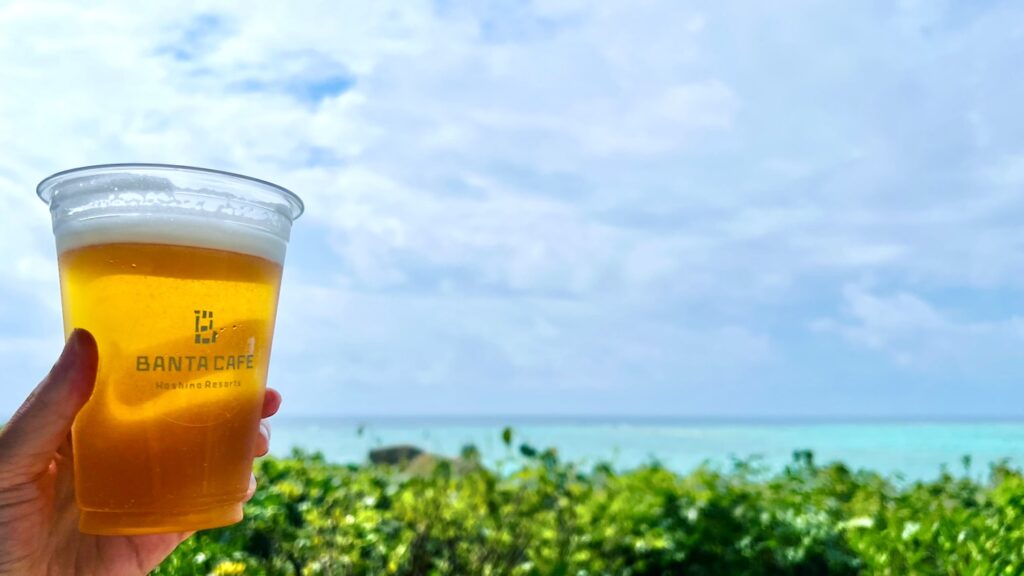 青空と青い海を背景に、BANTA CAFEのカップに入った生ビールを手に持っている風景
