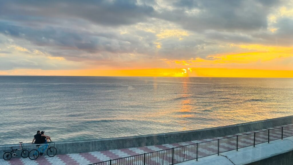 海に沈む綺麗な夕陽と宮城海岸の遊歩道と、防波堤に座り夕日を眺める2人と自転車がある風景