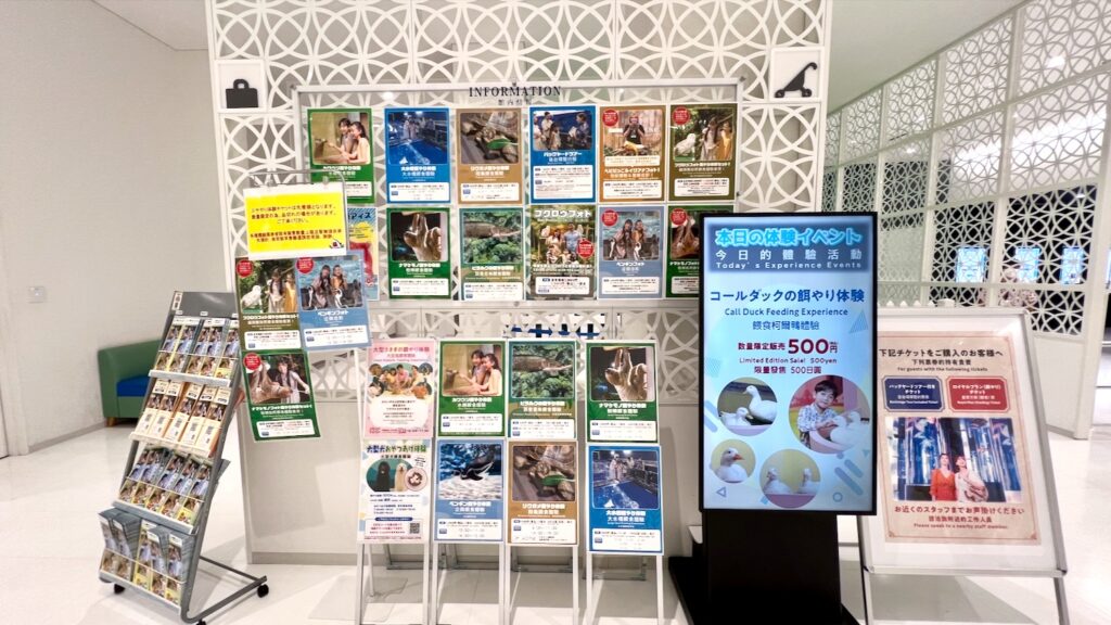 様々な動物との体験イベントのポスターが多数掲示されたインフォメーションボードを中心に、パンフレットラックや「本日の体験イベント」を表示するデジタルサイネージが並ぶ館内の案内エリア